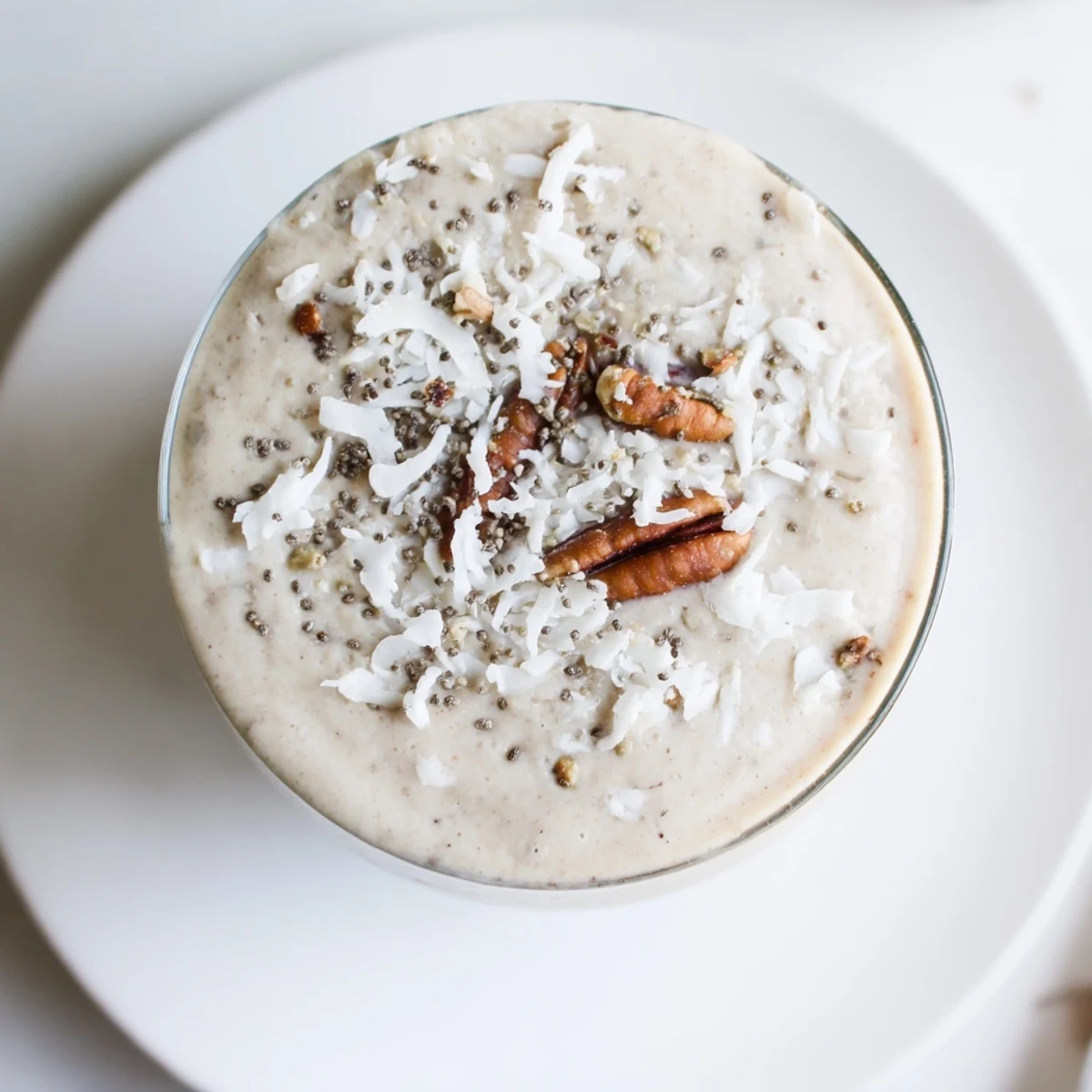 A glass of Snowy Day Paleo Smoothie topped with chia seeds and chopped nuts next to a cozy winter window.