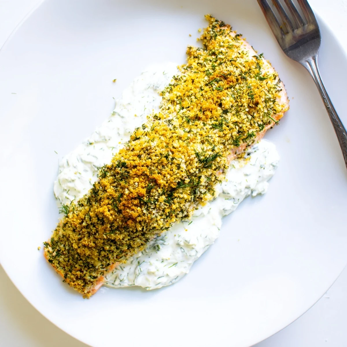 Fresh herb crusted salmon fillets with a golden topping, served alongside a white bowl of tangy dill sauce.
